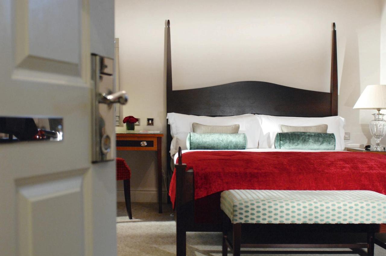 Macdonald Bath Spa Hotel Szoba fotó The photo shows a well-decorated hotel room. In the foreground, there is a partially opened door leading into the room. The focal point of the room is a large bed with a dark wooden headboard and stylish red and white bedding, accented with green pil