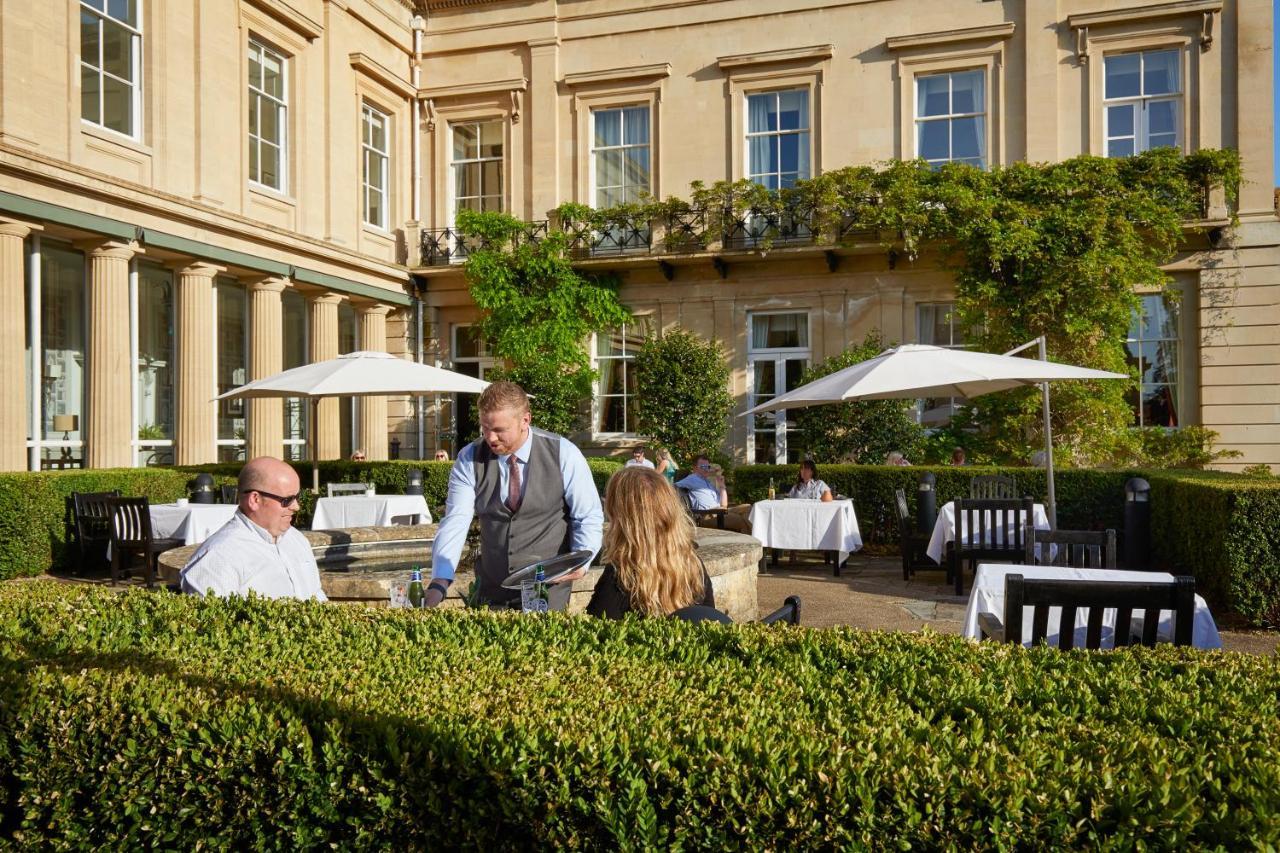 Macdonald Bath Spa Hotel Kültér fotó The photo depicts an outdoor dining area of a restaurant. In the foreground, there are neatly trimmed hedges surrounding the dining space. Two patrons are seated at a table; one is a man wearing sunglasses and the other is a woman facing him. A waite