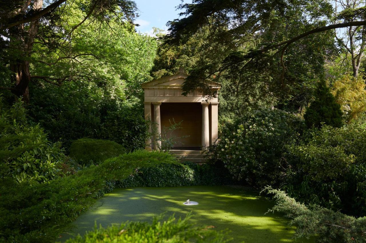 Macdonald Bath Spa Hotel Kültér fotó The photo shows a serene garden scene featuring lush greenery and a tranquil pond. In the background, there is a classical-looking structure with columns, resembling a small temple or pavilion. The pond appears to have vegetation covering its surface