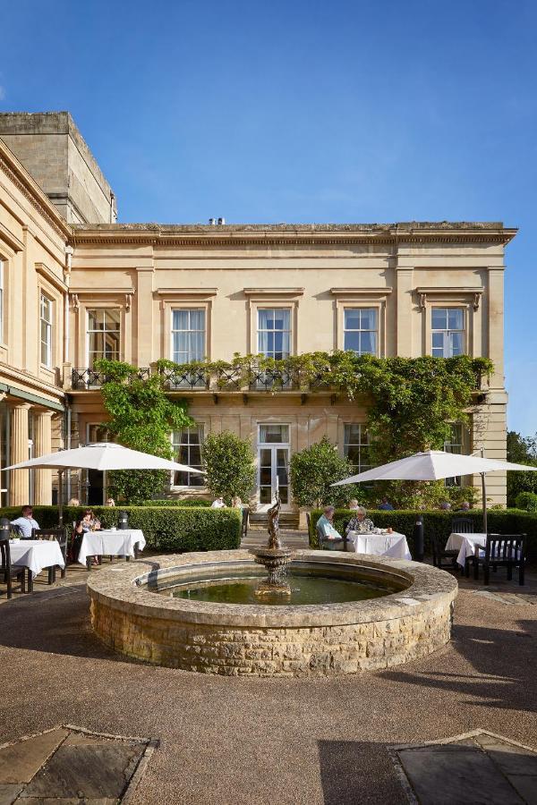 Macdonald Bath Spa Hotel Kültér fotó The photo depicts a charming outdoor dining area of a classic-looking building with elegant architecture. The structure features large windows and is adorned with greenery, including climbing plants. In the foreground, there is a circular fountain, a