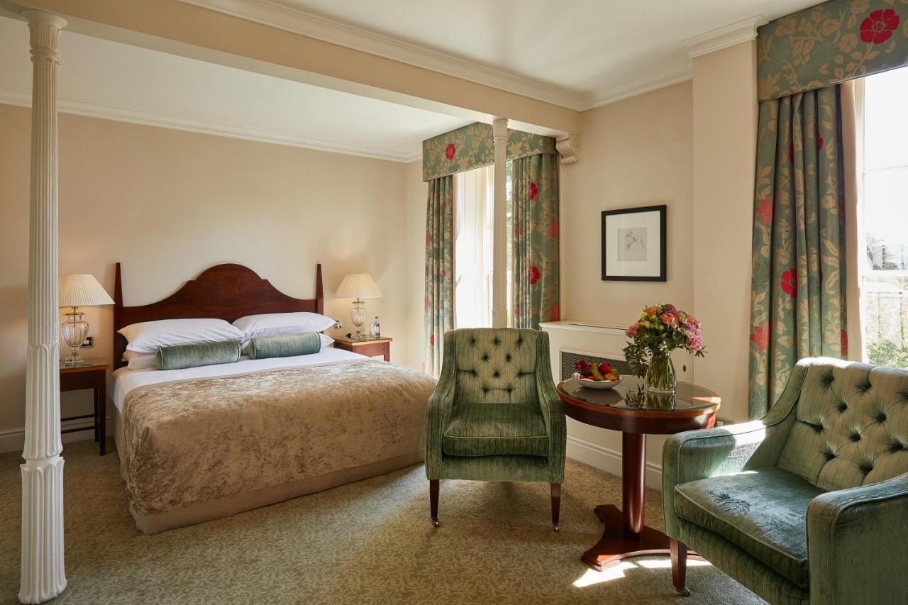 Macdonald Bath Spa Hotel Kültér fotó The photo shows a cozy hotel room featuring a large bed with a beige bedspread. There are two green armchairs positioned next to a small round table. The decor includes floral curtains and a neutral color palette on the walls. Soft lighting is provid