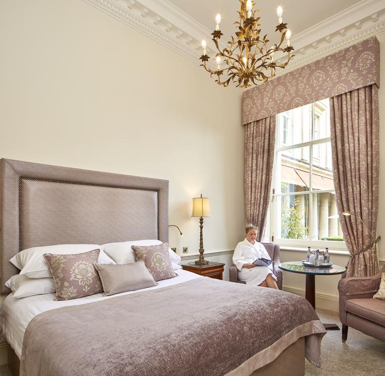 Macdonald Bath Spa Hotel Kültér fotó The photo shows a well-appointed hotel room that features a large, elegant bed with soft bedding and decorative pillows. The walls are painted a light color, and there is an ornate chandelier hanging from the ceiling. A beautiful window is adorned wi