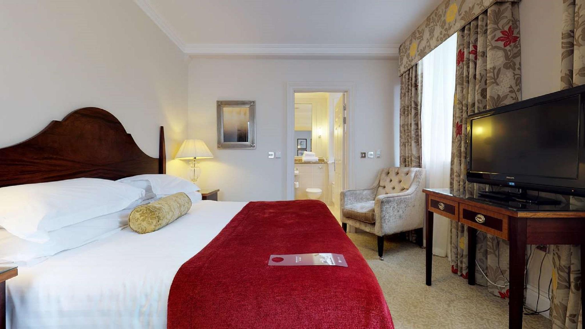 Macdonald Bath Spa Hotel Kültér fotó The photo shows a hotel room with a queen-sized bed covered in white linens and a deep red throw blanket. There is a decorative pillow on the bed. Next to the bed, there's a small nightstand with a lamp. In the background, a seating area features a b