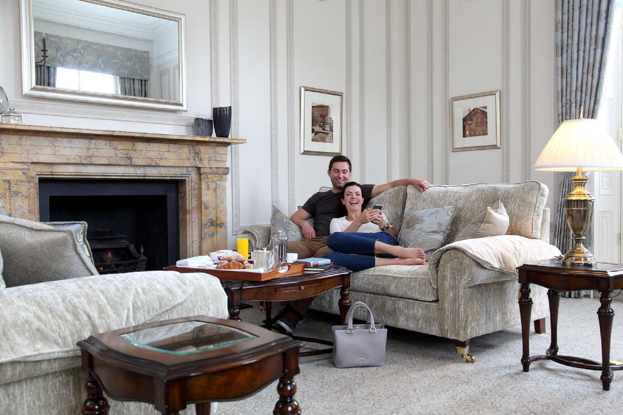 Macdonald Bath Spa Hotel Kültér fotó The photo shows a cozy living room setting where a couple is sitting together on a light-colored sofa. They appear relaxed, with the man sitting slightly behind the woman, who is smiling and looking at her phone. The room features elegant decor, incl