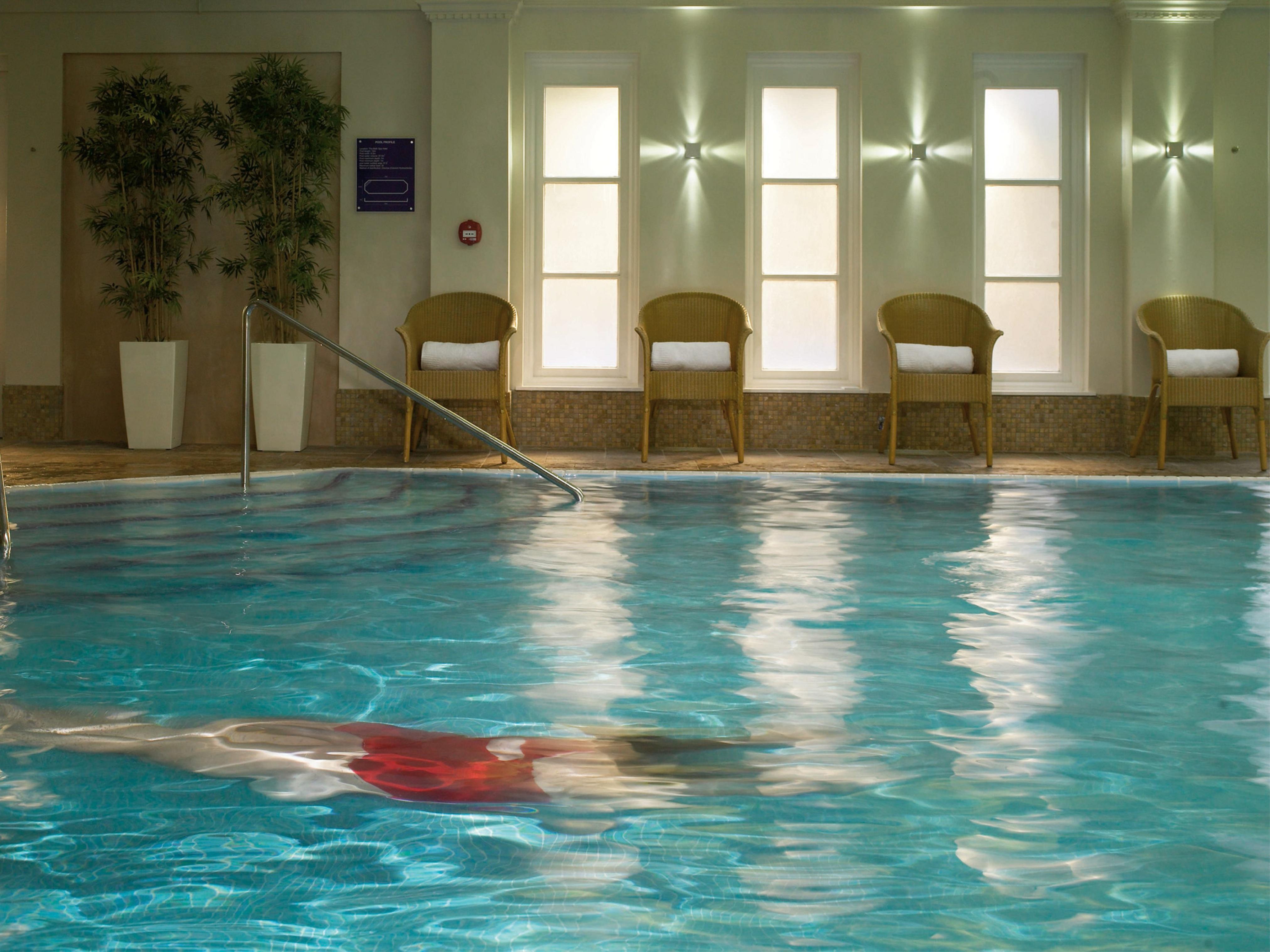Macdonald Bath Spa Hotel Kültér fotó The photo shows an indoor swimming pool with clear, blue water. In the pool, a person is swimming, partially submerged, and wearing a red swimsuit. Surrounding the pool, there are several empty chairs arranged along the side, along with potted plants