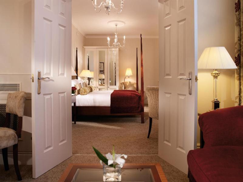 Macdonald Bath Spa Hotel Szoba fotó The photo shows a hotel room that can be viewed through open double doors. Inside, there is a neatly made bed with decorative pillows, a plush area rug, and soft lighting from lamps. The room features elegant furnishings, including a chair, a coffee 