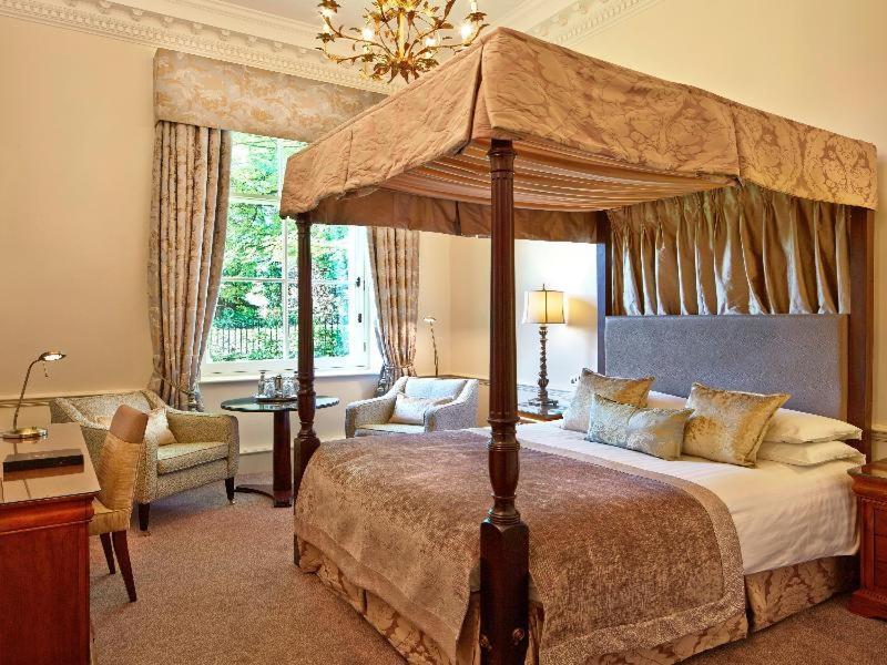 Macdonald Bath Spa Hotel Szoba fotó The photo shows an elegantly designed bedroom featuring a four-poster bed with a decorative canopy. The bed is dressed with luxurious bedding and pillows, creating a cozy and inviting atmosphere. Flanking the bed are two comfortable armchairs and a s