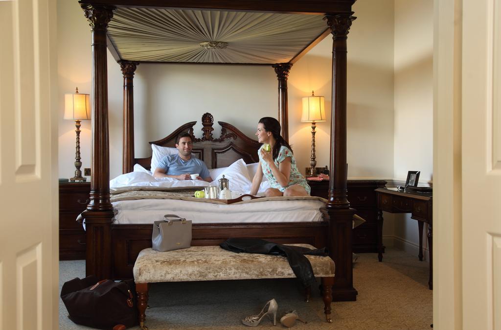 Macdonald Bath Spa Hotel Kültér fotó The image depicts a spacious, well-lit bedroom featuring a large four-poster bed with an ornate headboard. A man and a woman are sitting on the bed, appearing to engage in conversation. The woman is dressed in a patterned outfit, while the man is wea